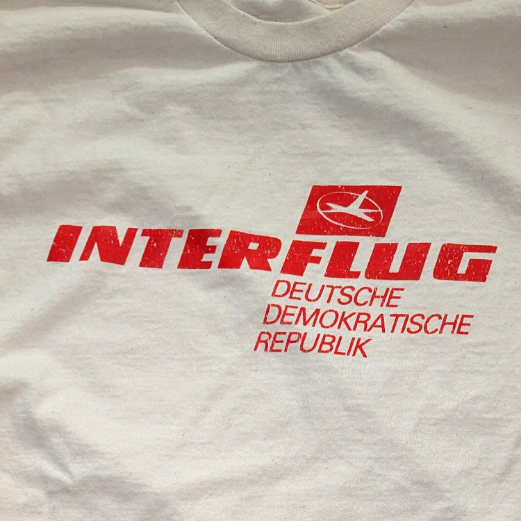 Close-up of a white t-shirt featuring a red distressed graphic print with the text 'INTERFLUG' and 'DEUTSCHE DEMOKRATISCHE REPUBLIK' alongside a small airplane logo.