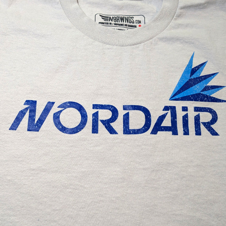 Close-up of a light heather gray crewneck t-shirt featuring a distressed blue 'NORDAIR' logo and a blue stylized bird graphic on the chest. The inner collar label shows 'NORDWINGS.