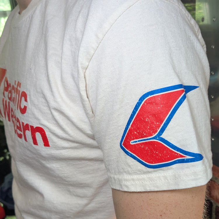 Close-up of a light beige or off-white t-shirt featuring a distressed red graphic of the Pacific Western airline logo on shoulder