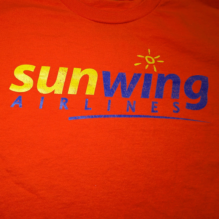 Orange crewneck t-shirt featuring the 'sunwing AIRLINES' logo printed across the chest in yellow and dark blue, with small yellow sun graphics on both sleeves closeup.