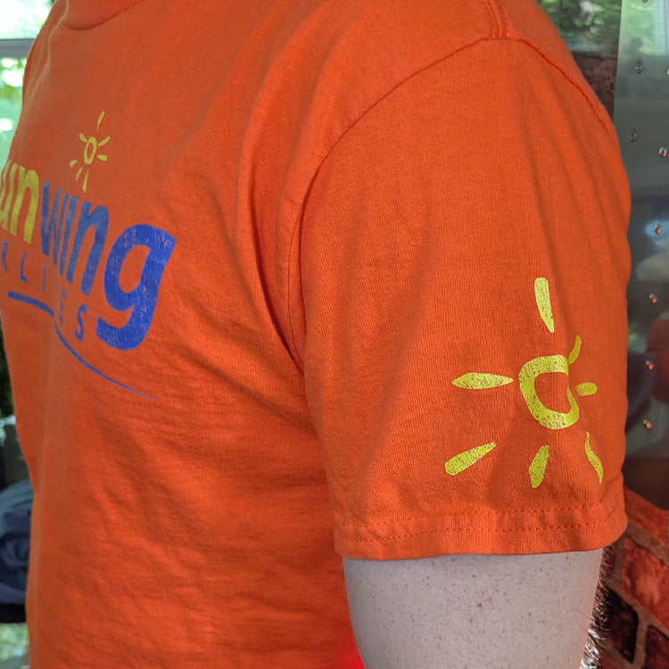 Orange crewneck t-shirt featuring the 'sunwing AIRLINES' logo printed across the chest in yellow and dark blue, with small yellow sun graphics on both sleeves closeup.